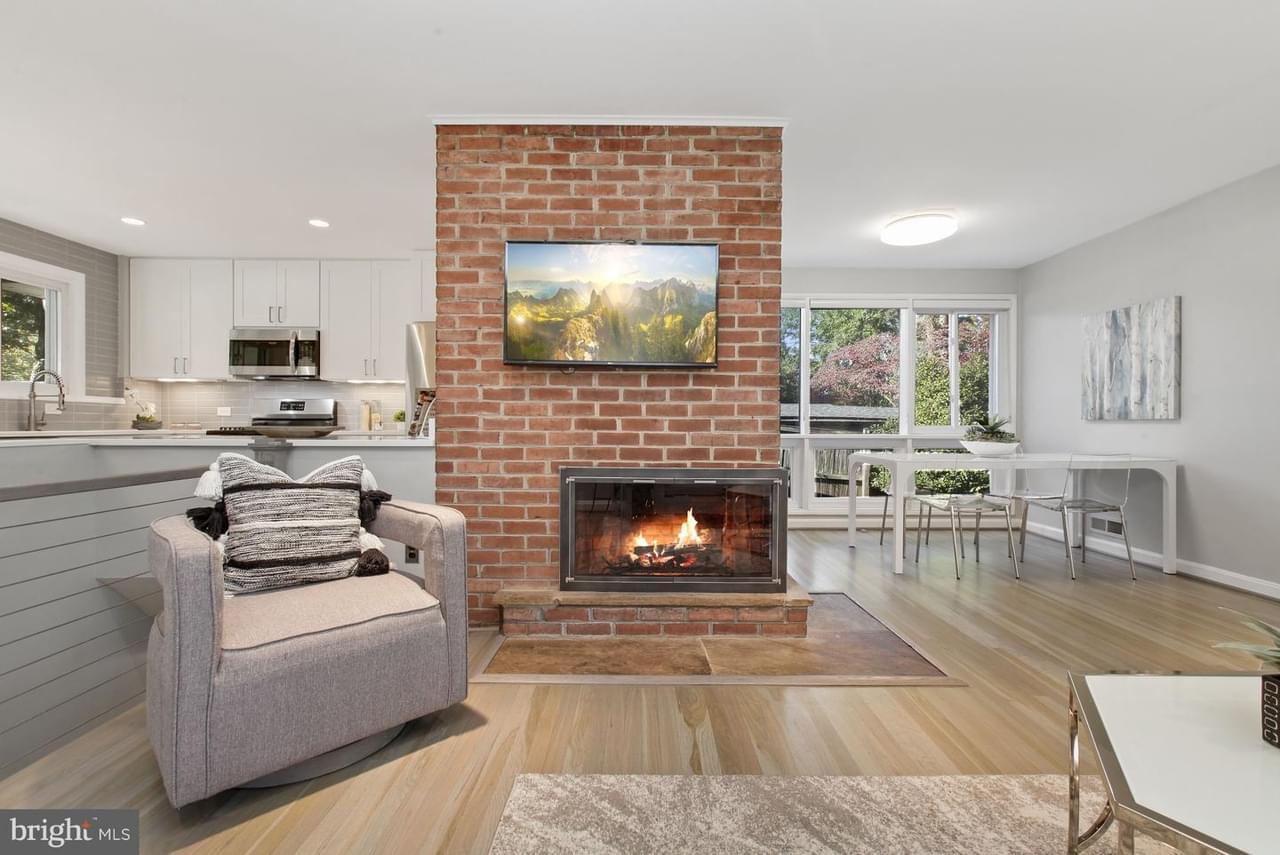 A light colored open concept area with light colored wood floor, brick fireplace with TV over the top, view to the kitchen and dining area, large windows